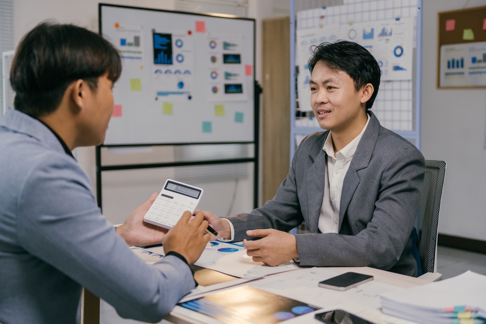 Two young asian businessmen are discussing company performance using a calculator and financial reports with charts and graphs, analyzing data and planning strategies for growth and success